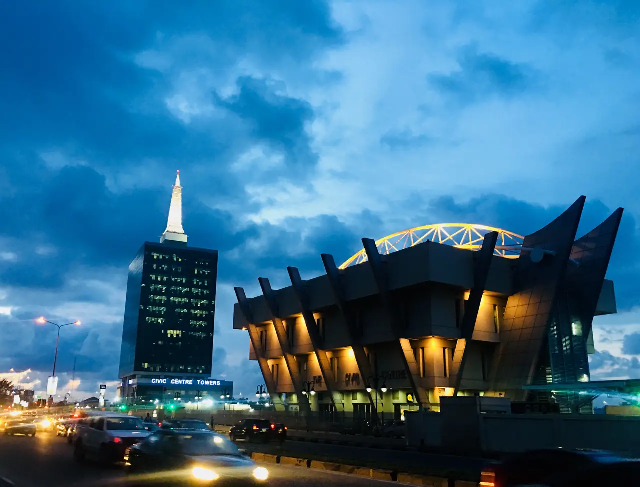 The Civic Centre and Civic Towers lit up at dusk beside Ozumba Mbadiwe Avenue, Victoria Island, Lagos, with cars passing on the road.