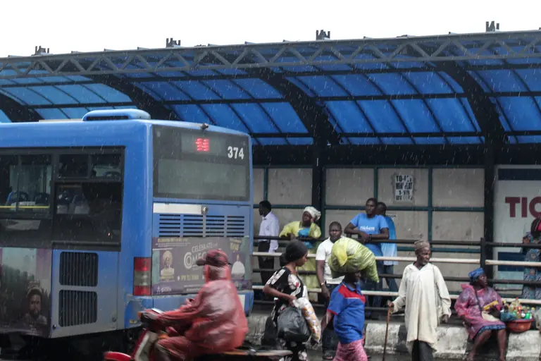 Passengers wait under the blue canopy of the Mile 12 BRT terminal in Lagos as a bus pulls in beside the ticketing area.